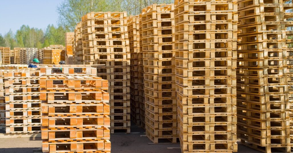 Several tall stacks of lightly colored wooden pallets outside of a warehouse. Two workers stand in the background.