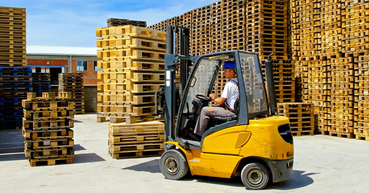 A man driving a small yellow forklift through an outdoor storage area with several tall stacks of wooden pallets.