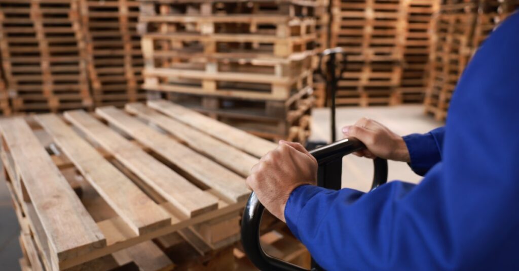 A person wearing long blue sleeves is using a pallet jack to move a stack of wooden pallets around a warehouse full of pallets.