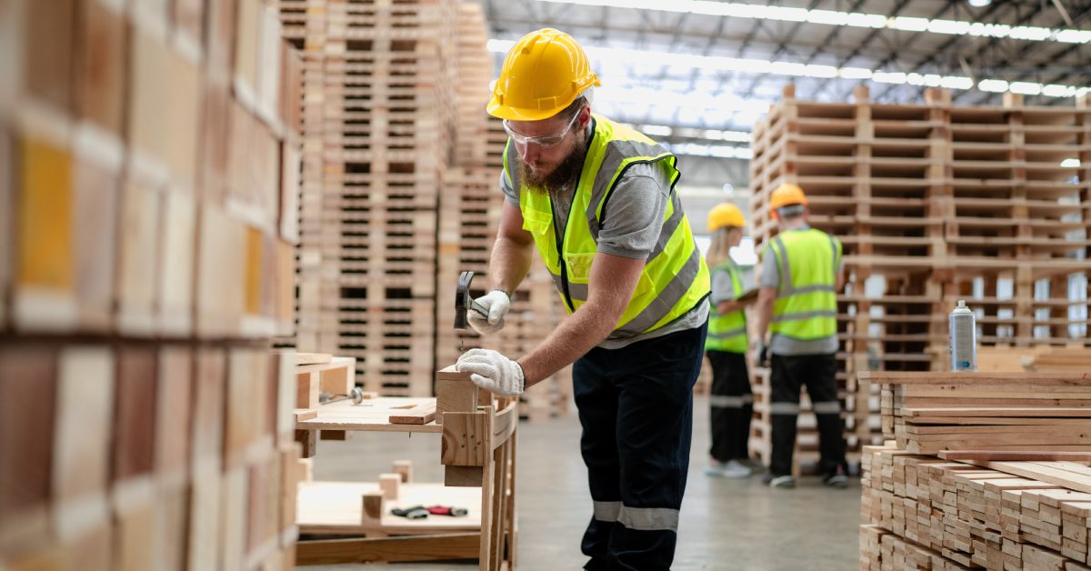 A man wearing PPE is standing in a warehouse full of pallets. He's using a hammer and nail to work on one of them.
