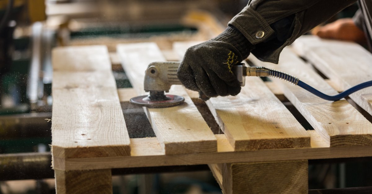 A gloved hand is holding a small power sander. It's using the rounded disc on the surface of a wood pallet.
