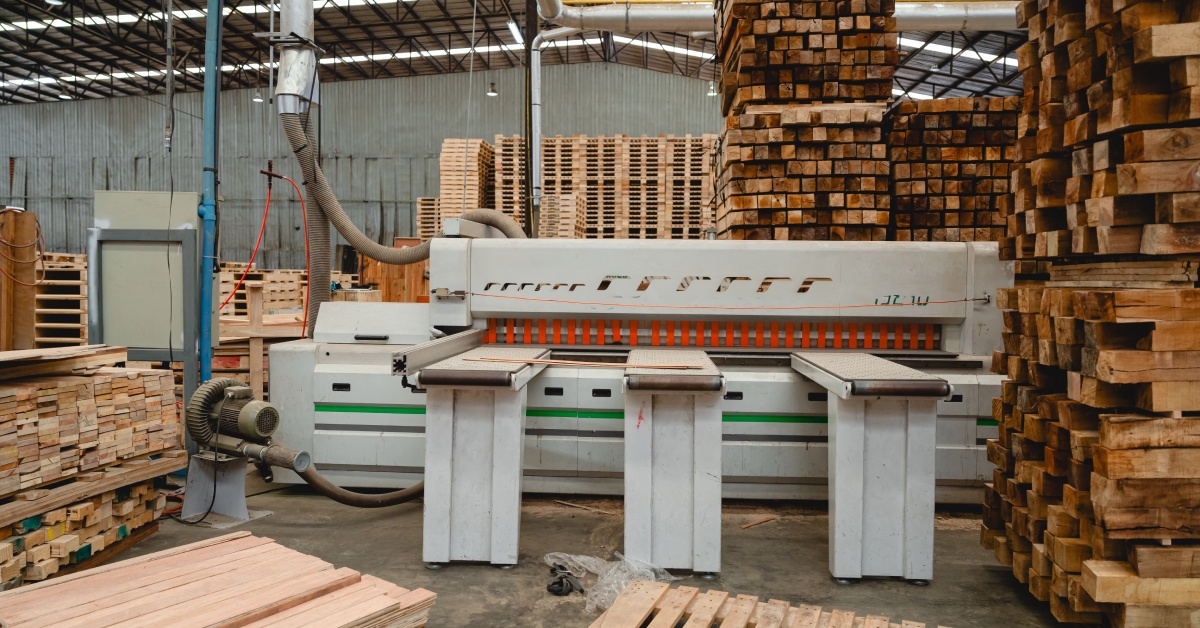 A warehouse with stacks of wooden pallets, planks of wood, and a machine placed in the middle of the facility for cutting wood.